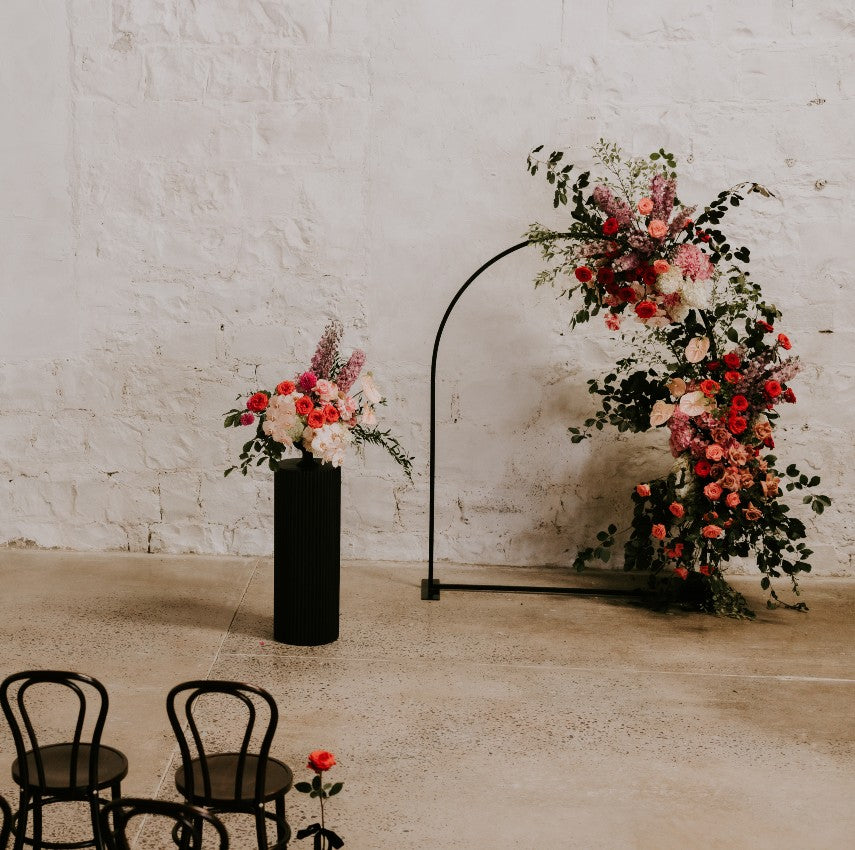 Floral arch and vase against a white stone wall with two brown chairs.