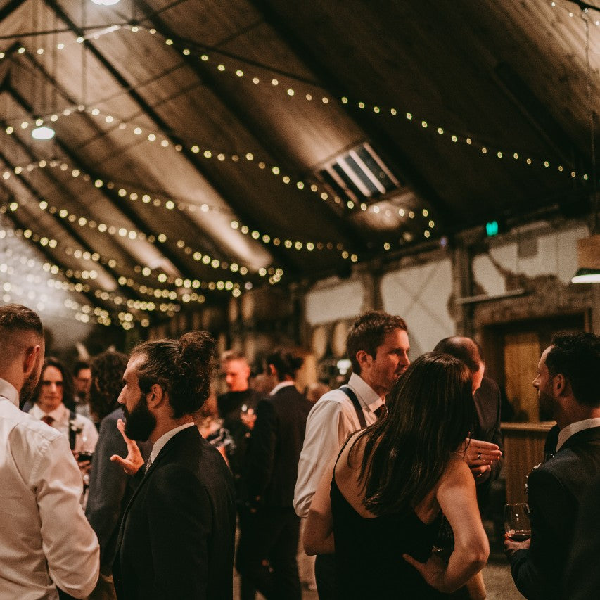 Under festoon lighting people dance and chat at a function in a winery barrel hall