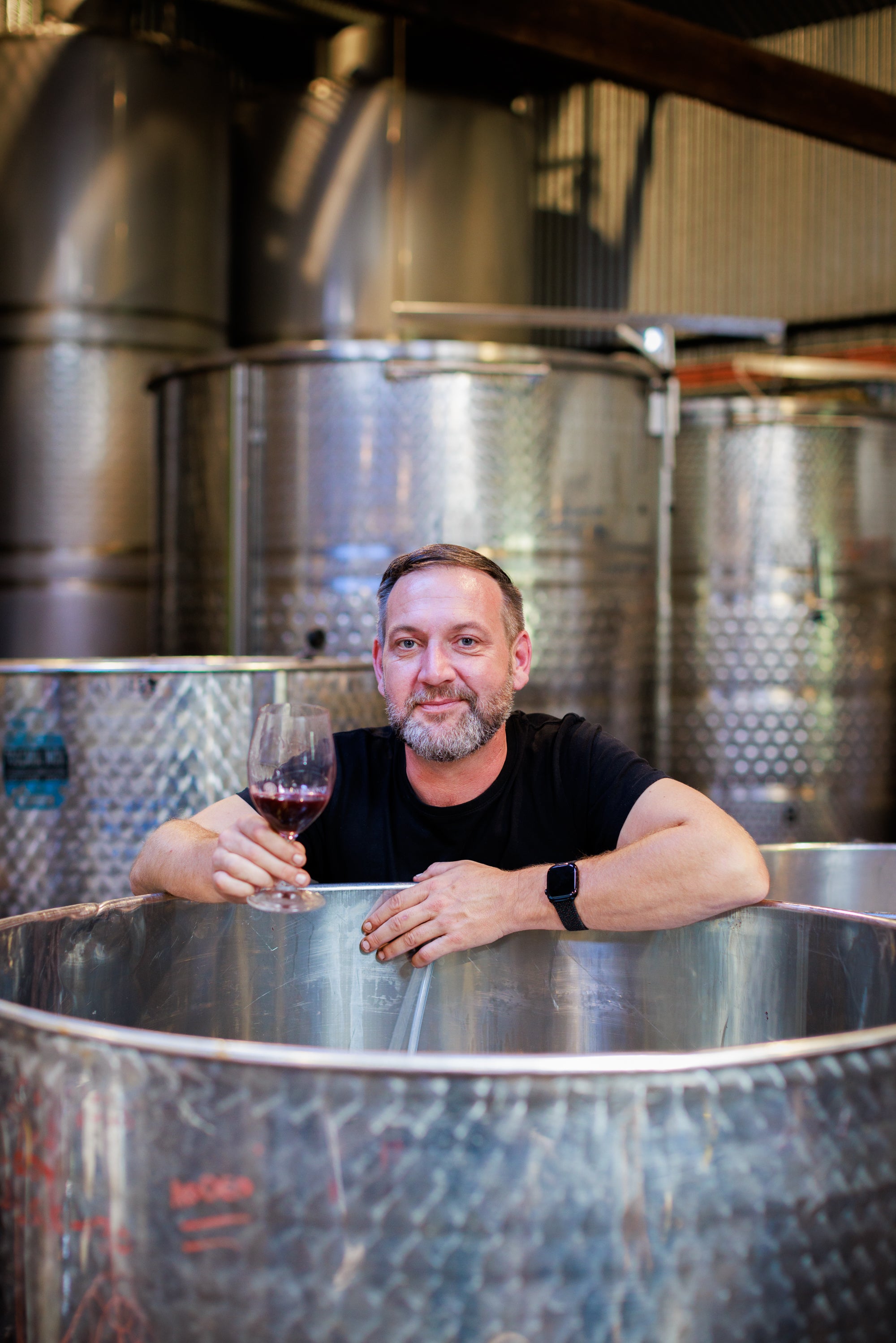 Man holding a glass of red wine in front of large metal wine tanks.