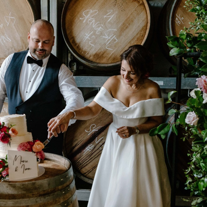 Couple cutting a wedding cake in front of wine barrels with floral decorations.