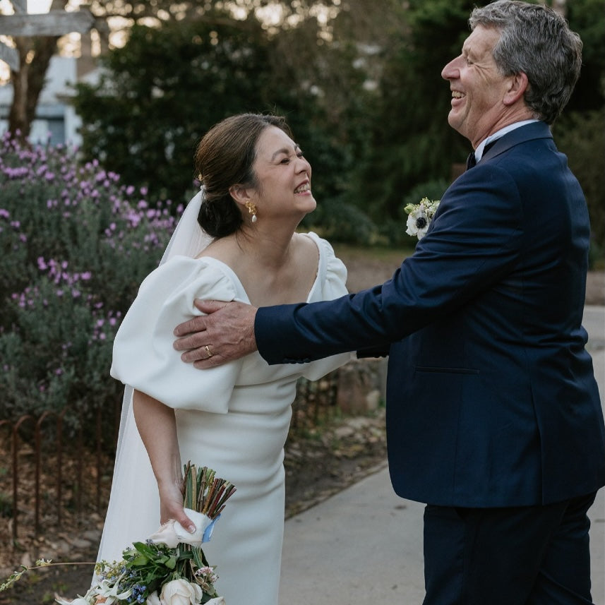 Man and woman in wedding attire standing outdoors with a bouquet and floral arrangement.